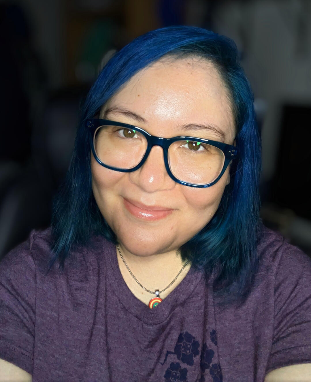 Portrait of Amy A. Cowan a woman with blue hair and big, blue glasses smiles at the camera
